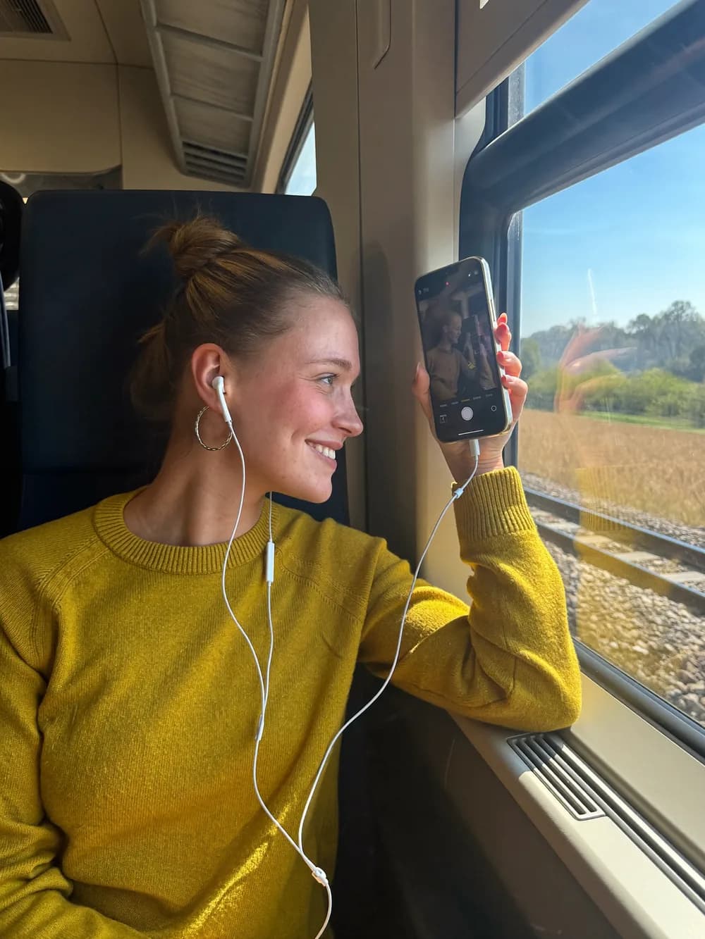 Ragazza col maglione giallo che sorride al telefono, seduta vicino al finestrino del treno